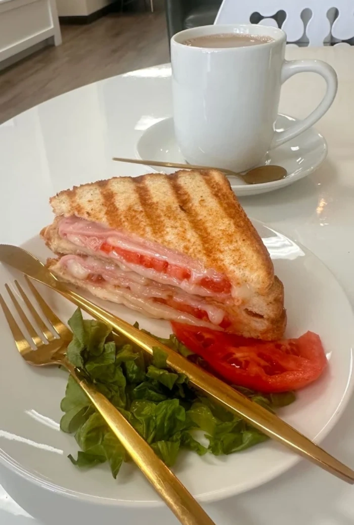 Grilled ham and cheese sandwich with tomato, served with a side of lettuce and a cup of hot cocoa.