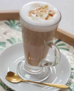 Café latte servido en vaso de cristal con espuma de leche y caramelo sobre plato blanco.